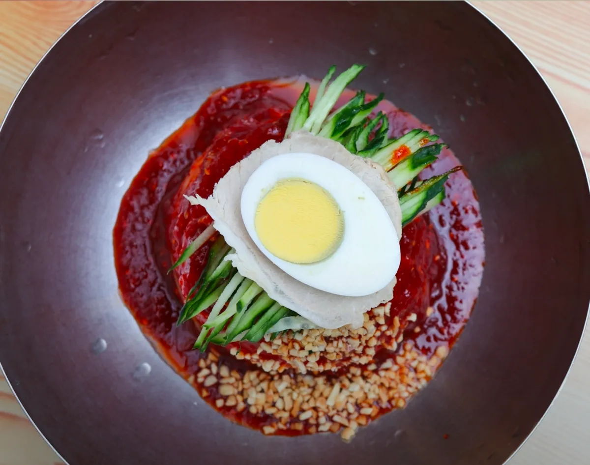 Overhead view of bibim milmyeon in a steel bowl with red sauce, sliced pork, boiled egg, cucumber and crushed peanuts