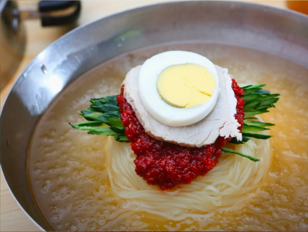 Mul milmyeon in a steel bowl of cold broth topped with red sauce, sliced pork, half a boiled egg and cucumber