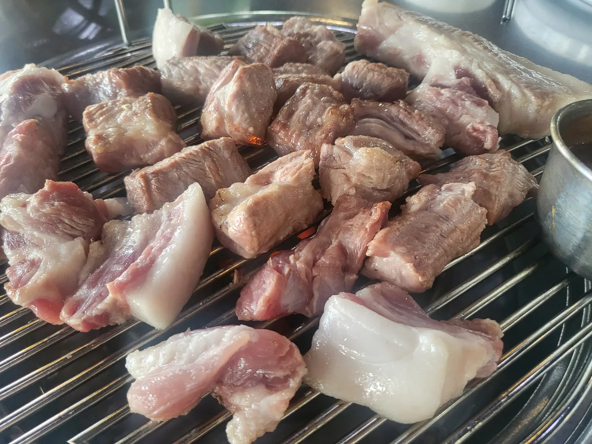 Pieces of pork on a round wire grill with a small steel cup of dipping sauce at the right edge