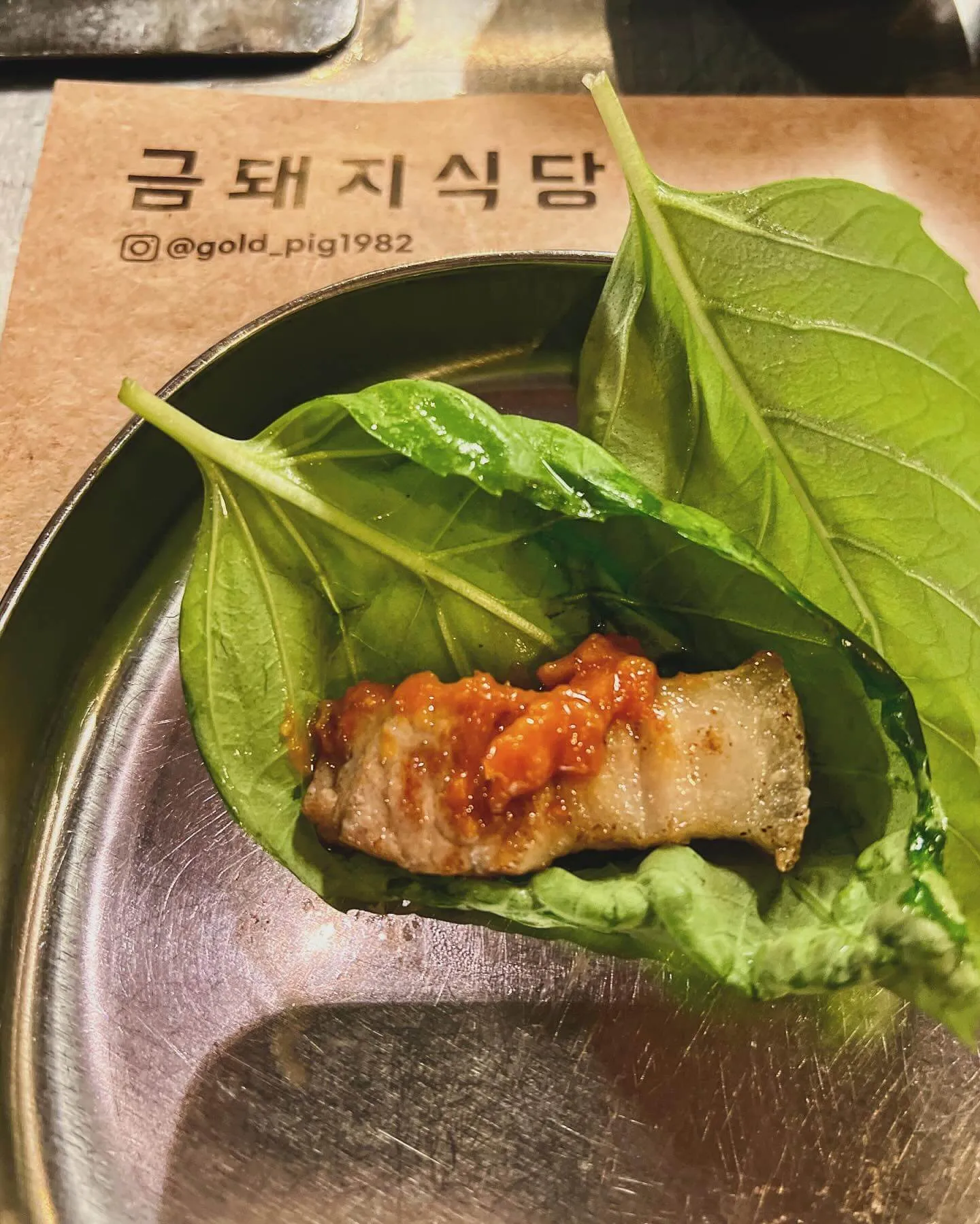 A slice of grilled pork with sauce on ssam leaves, on a steel plate at Geumdwaeji Sikdang