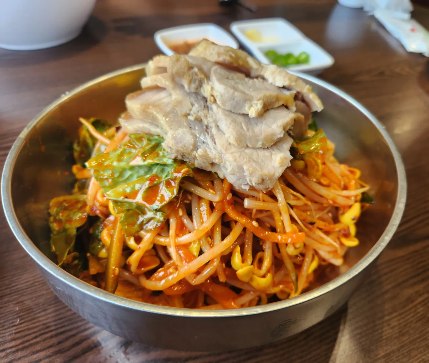 Steel bowl of red bibim noodles with bean sprouts, greens and sliced boiled pork on top