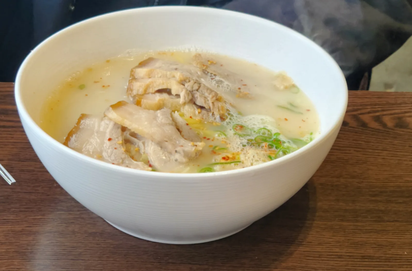 White bowl of pale pork noodle soup with sliced boiled pork, scallions and chili flakes