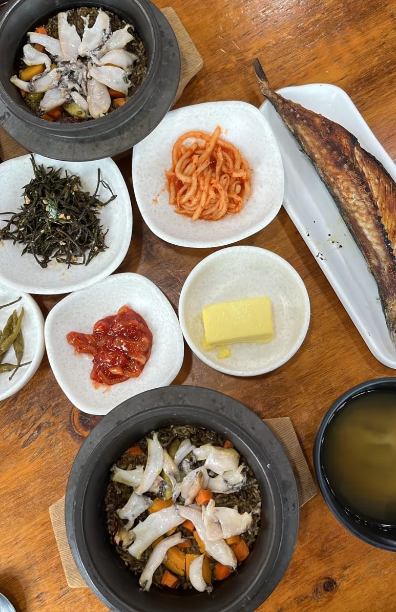 Overhead view of two abalone rice stone pots, a grilled mackerel, a butter pat and small side dishes on a wooden table