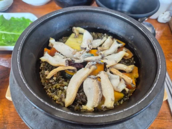 A black stone pot of rice topped with sliced abalone, pumpkin and carrot, with a green leaf side dish at the left edge