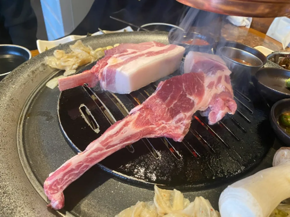 Raw pork cuts with a bone-in rib piece on a round grill, with steel sauce cups and sliced onion around it