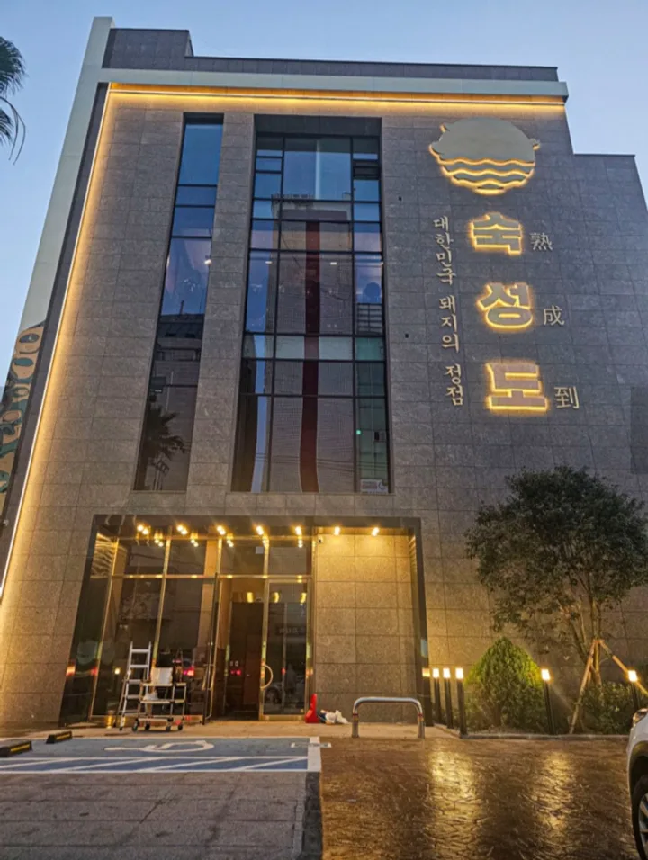 Stone-clad multi-story building with the 숙성도 name and a pig logo lit on the upper facade at dusk