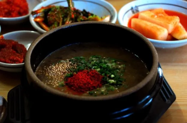 Black earthenware bowl of soup topped with scallions, sesame seeds and chili flakes, with kimchi side dishes behind