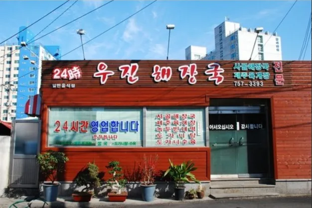 Red-paneled single-story restaurant front with a Korean sign, potted plants and apartment towers behind