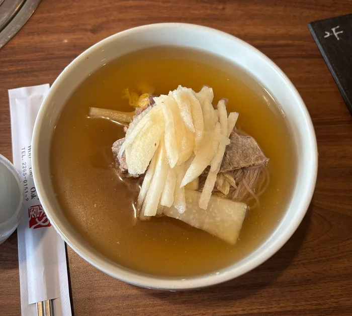 A white bowl of Pyongyang naengmyeon in clear broth with sliced pear, brisket, and buckwheat noodles, chopstick sleeve at the left edge
